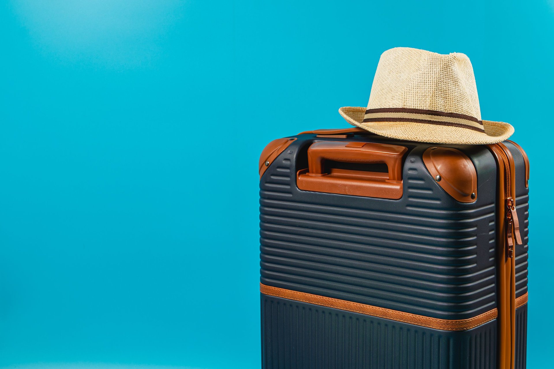 A stylish suitcase rests against a vibrant blue backdrop, topped with a straw hat, suggesting readiness for summer travel adventures