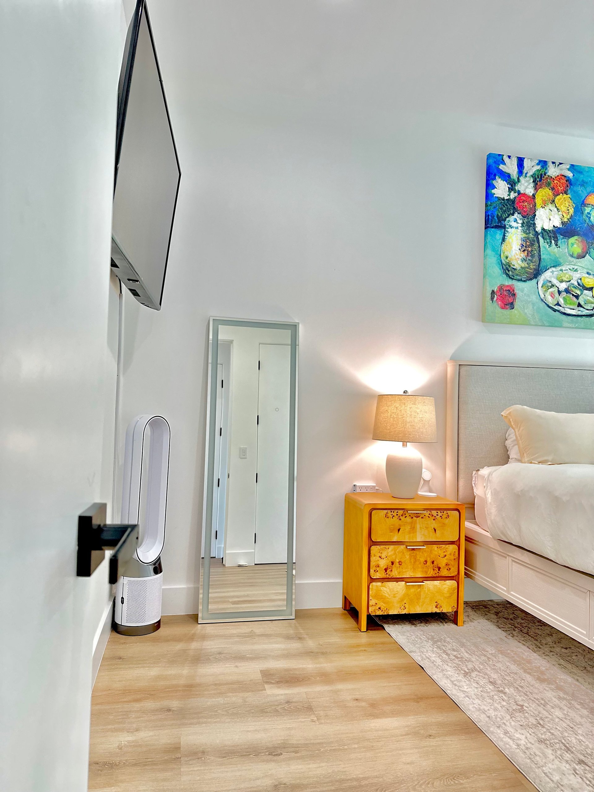 Modern bedroom with white bed, yellow nightstand, table lamp, framed artwork, and wooden flooring