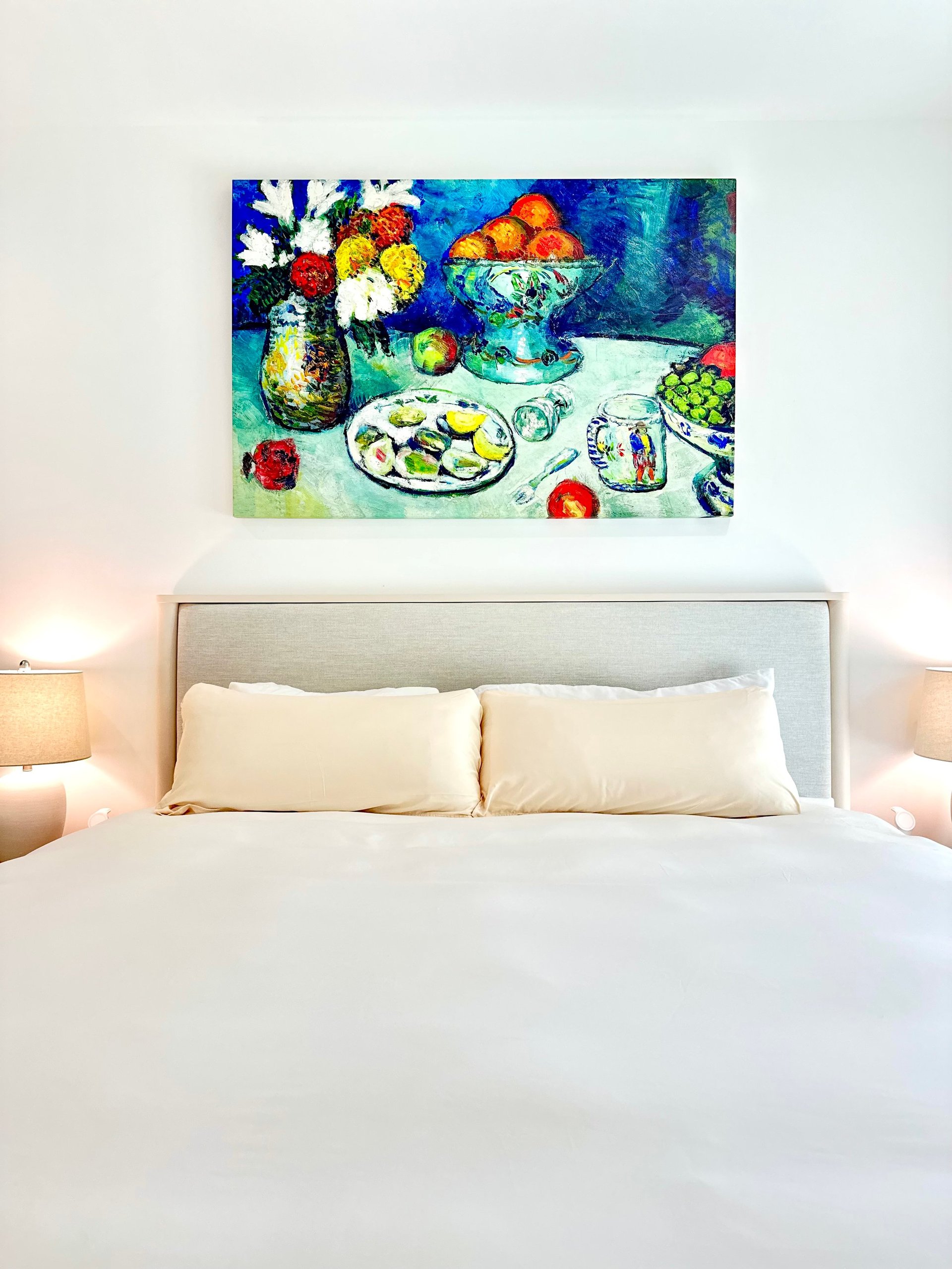 Colorful still life painting of flowers, fruits, and plates hanging above a bed with white bedding and neutral headboard