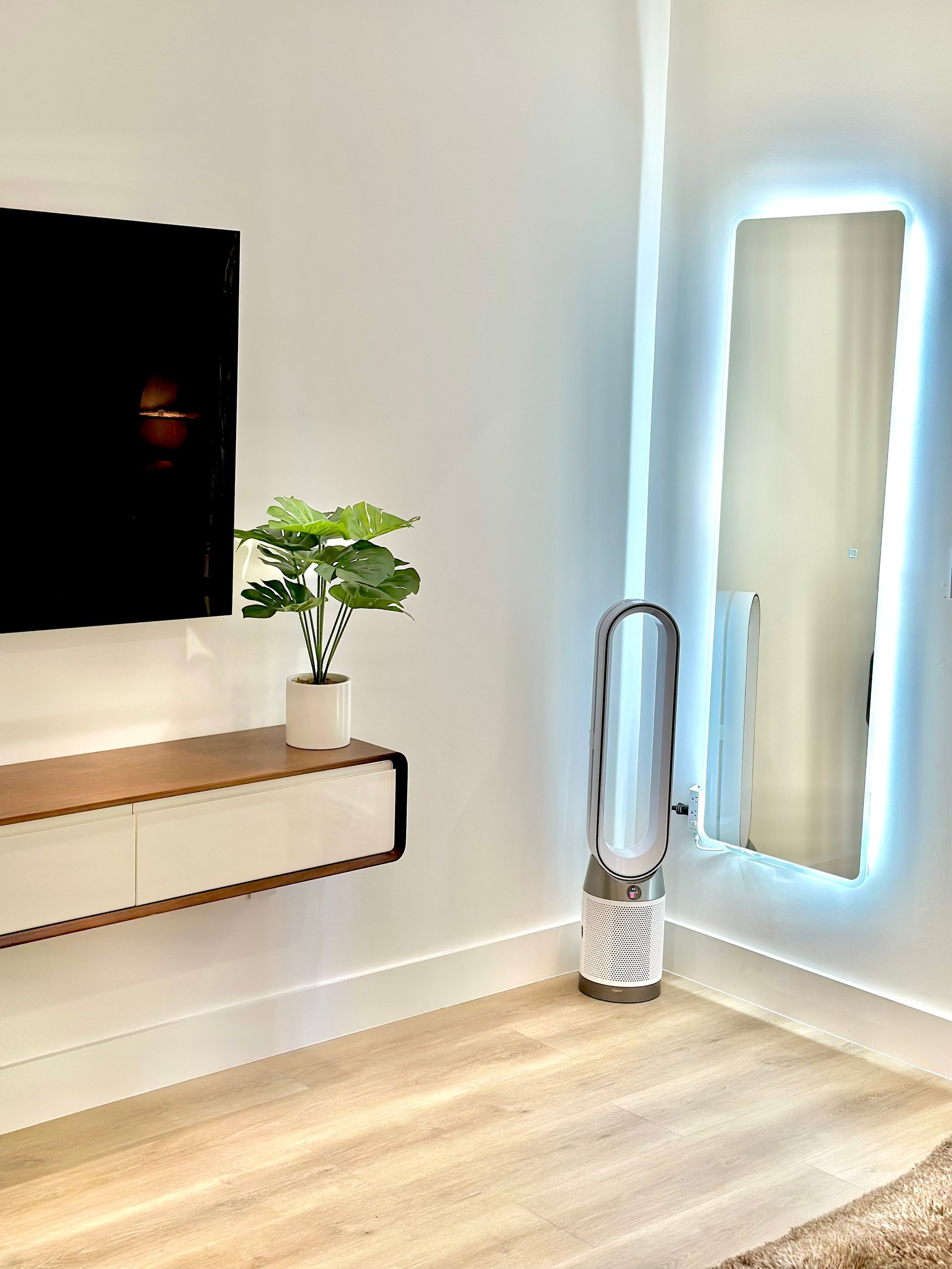 Modern living room with wall-mounted black TV, white console table with potted plant, and illuminated blue-trimmed mirror