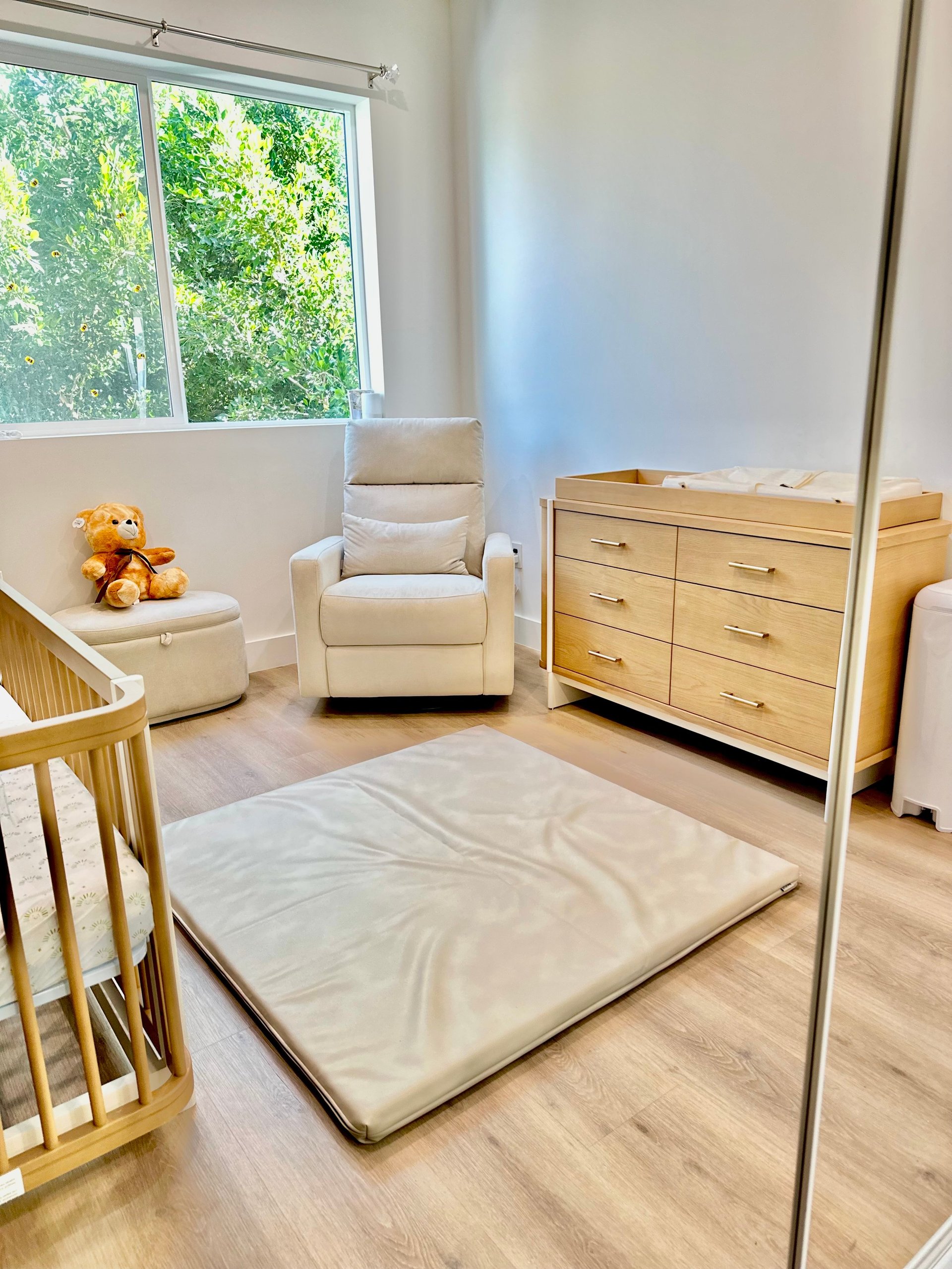Bright modern nursery room with wooden crib, cream armchair, wooden dresser, play mat, and large windows overlooking trees