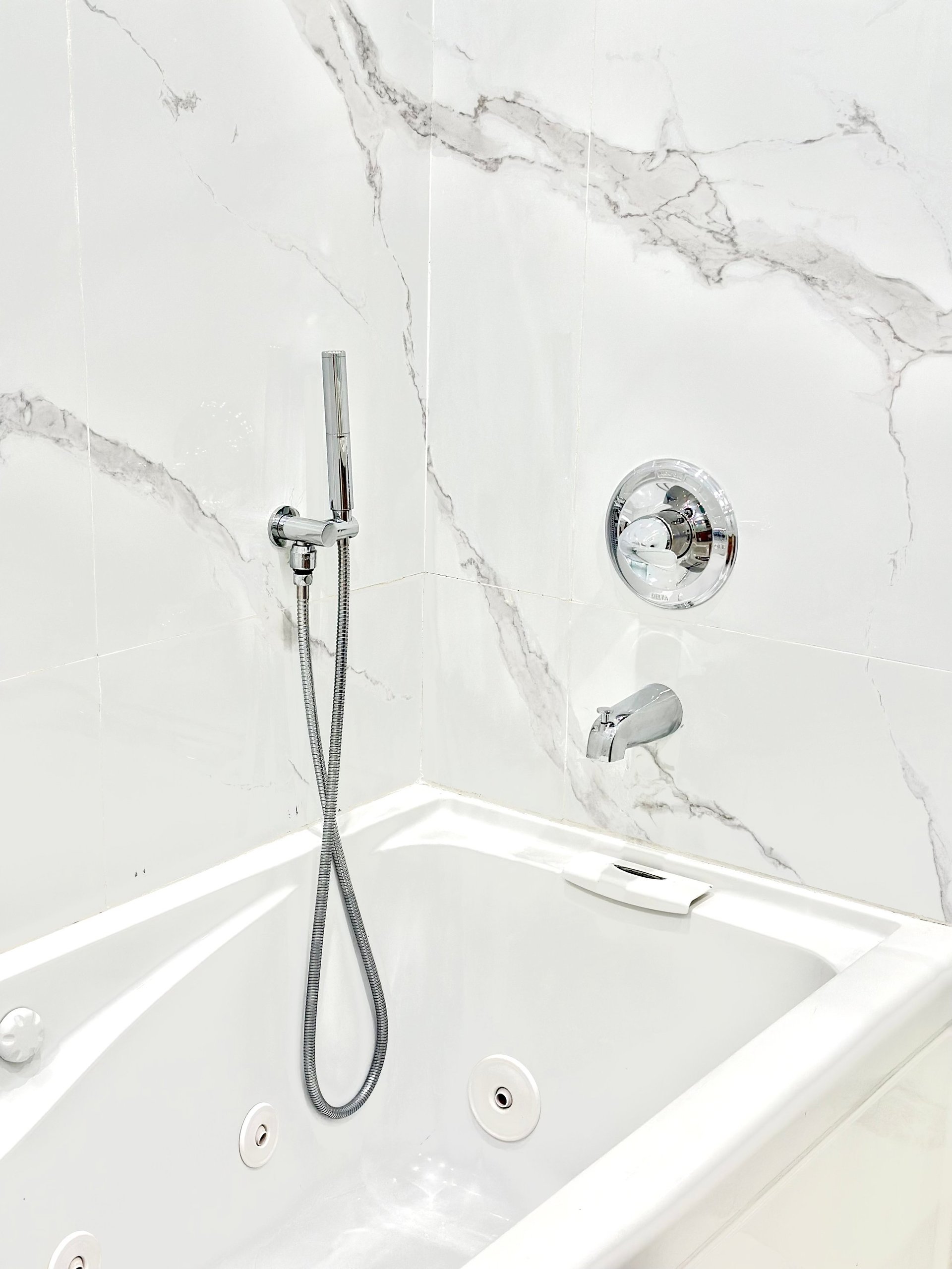 Modern white bathtub with chrome shower fixtures and marble-tiled surround wall