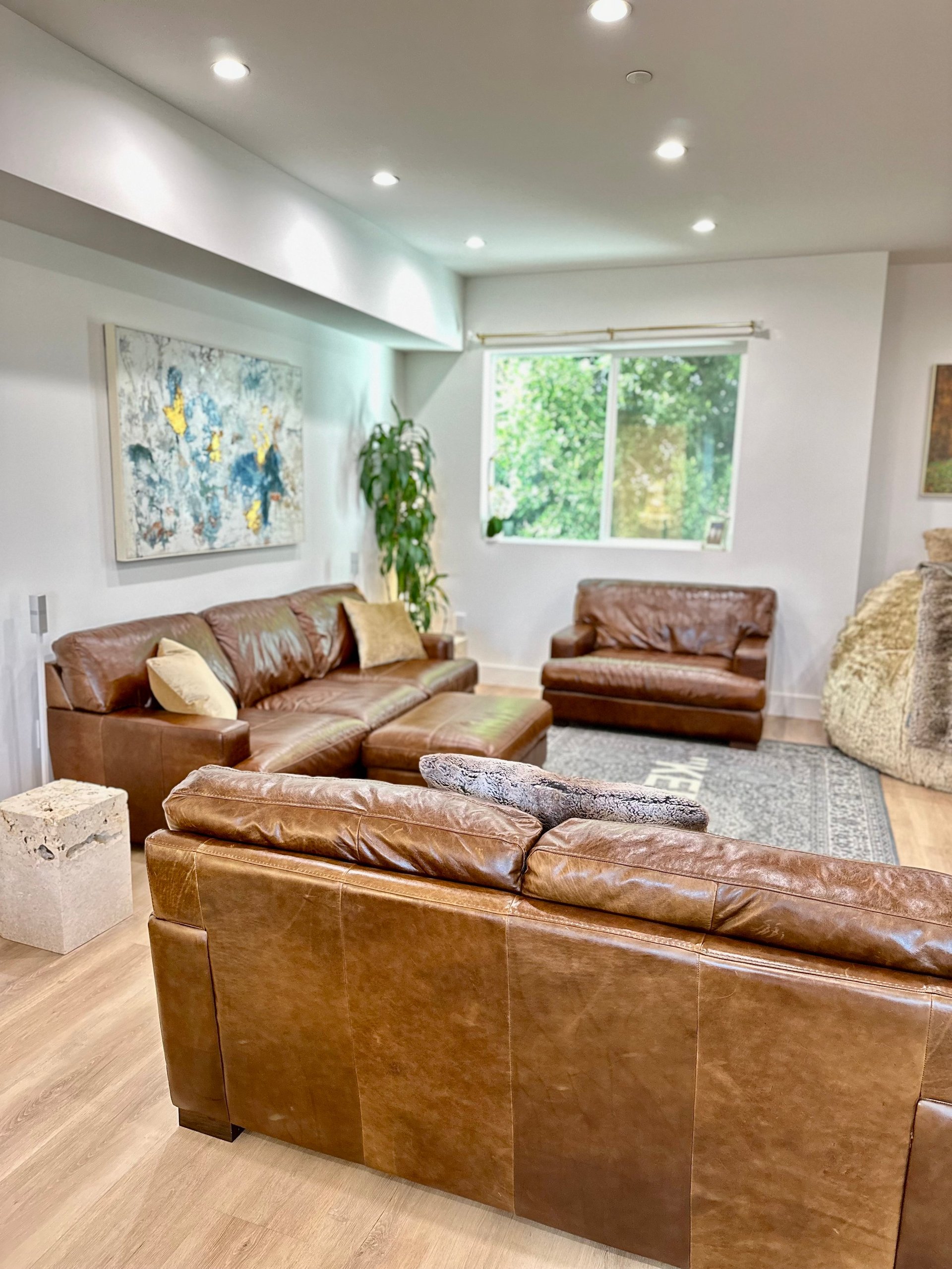 Modern living room with three brown leather sofas, recessed ceiling lights, large windows with greenery view, blue abstract wall art, and light wood flooring