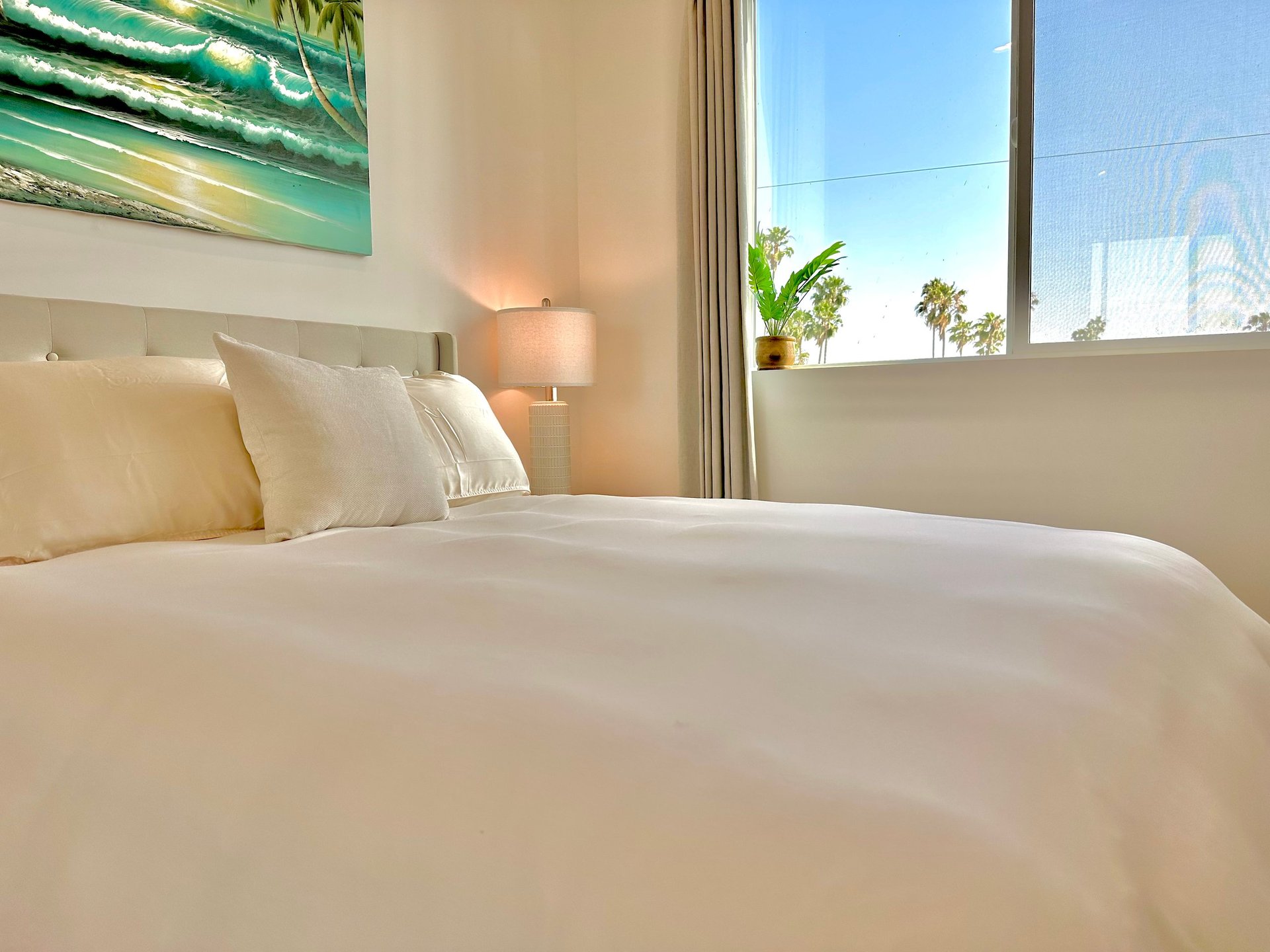 Modern bedroom with white bed, cream bedding, pillows, side lamp, and large windows overlooking palm trees and cityscape