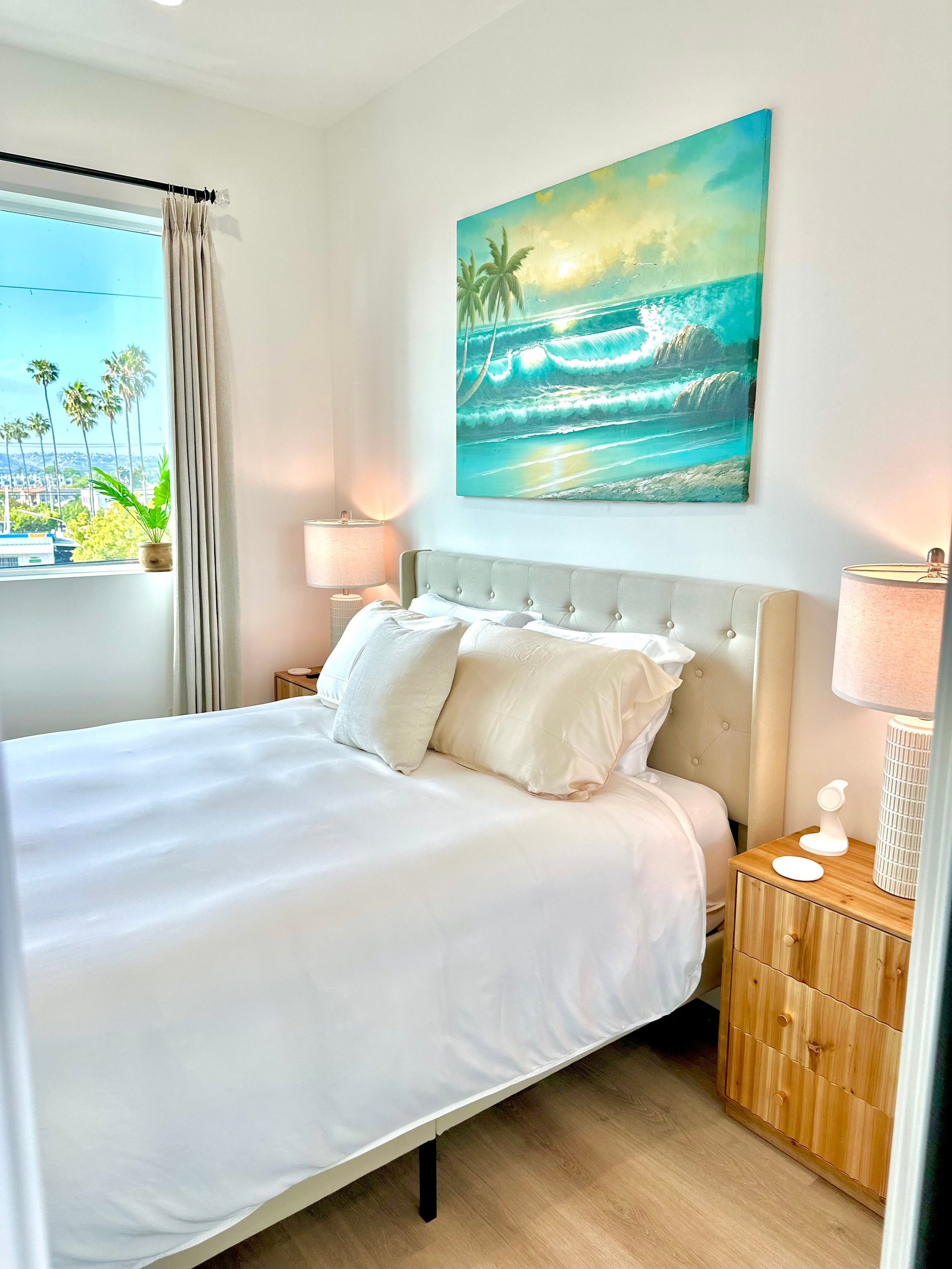 Bright bedroom with white bed, beige headboard, tropical wave artwork above bed, wooden nightstands, and window with palm trees visible outside.