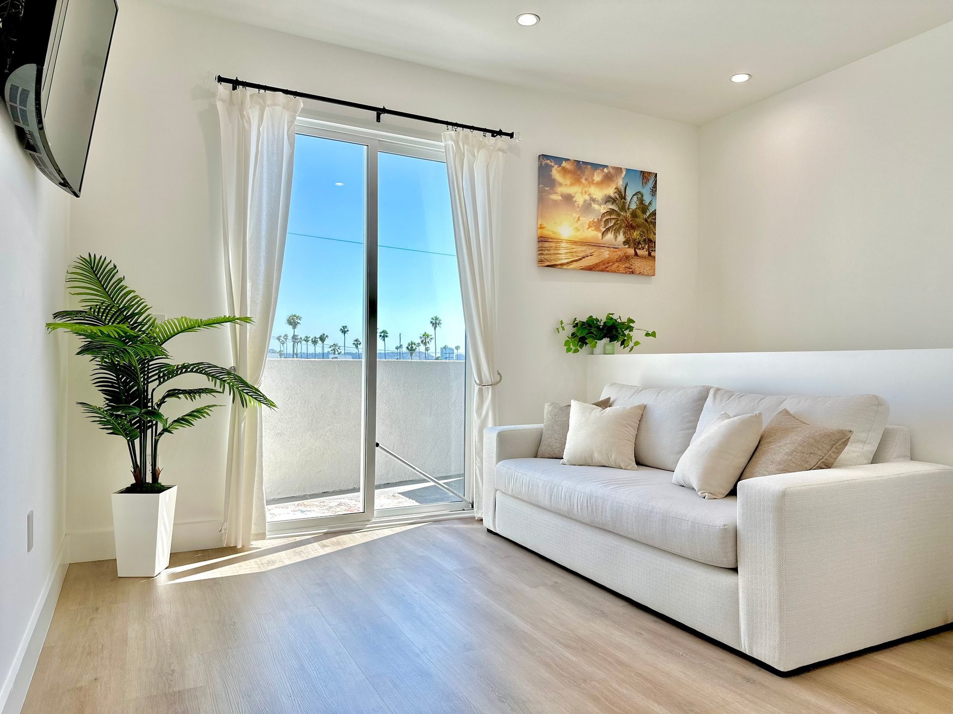 Modern living room with white sofa, potted plant, sliding glass doors with palm tree view, and wooden flooring