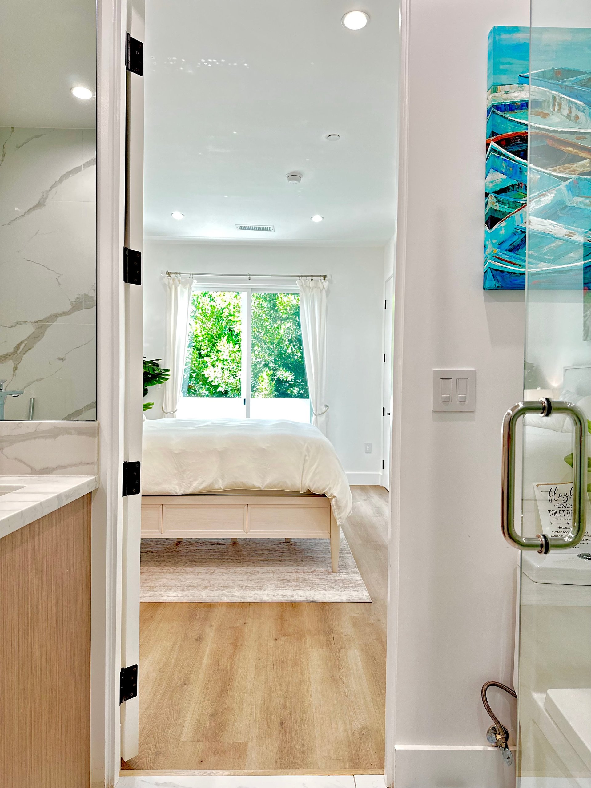Bright, modern master bedroom viewed through an open doorway from a bathroom, featuring a wood-frame bed, windows, and hardwood flooring