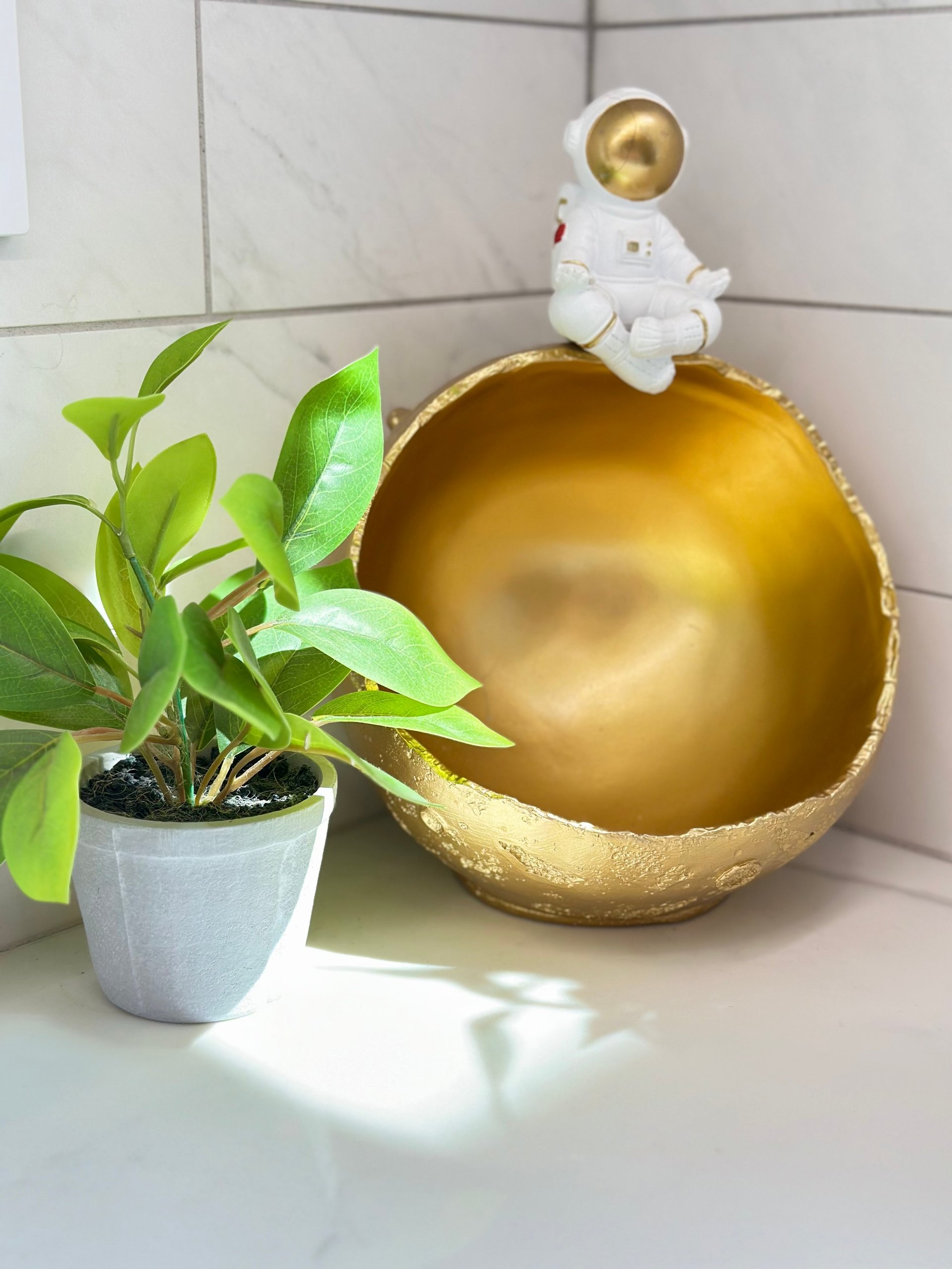 White astronaut figurine sitting on a large gold sphere planter next to a potted green plant on a shelf