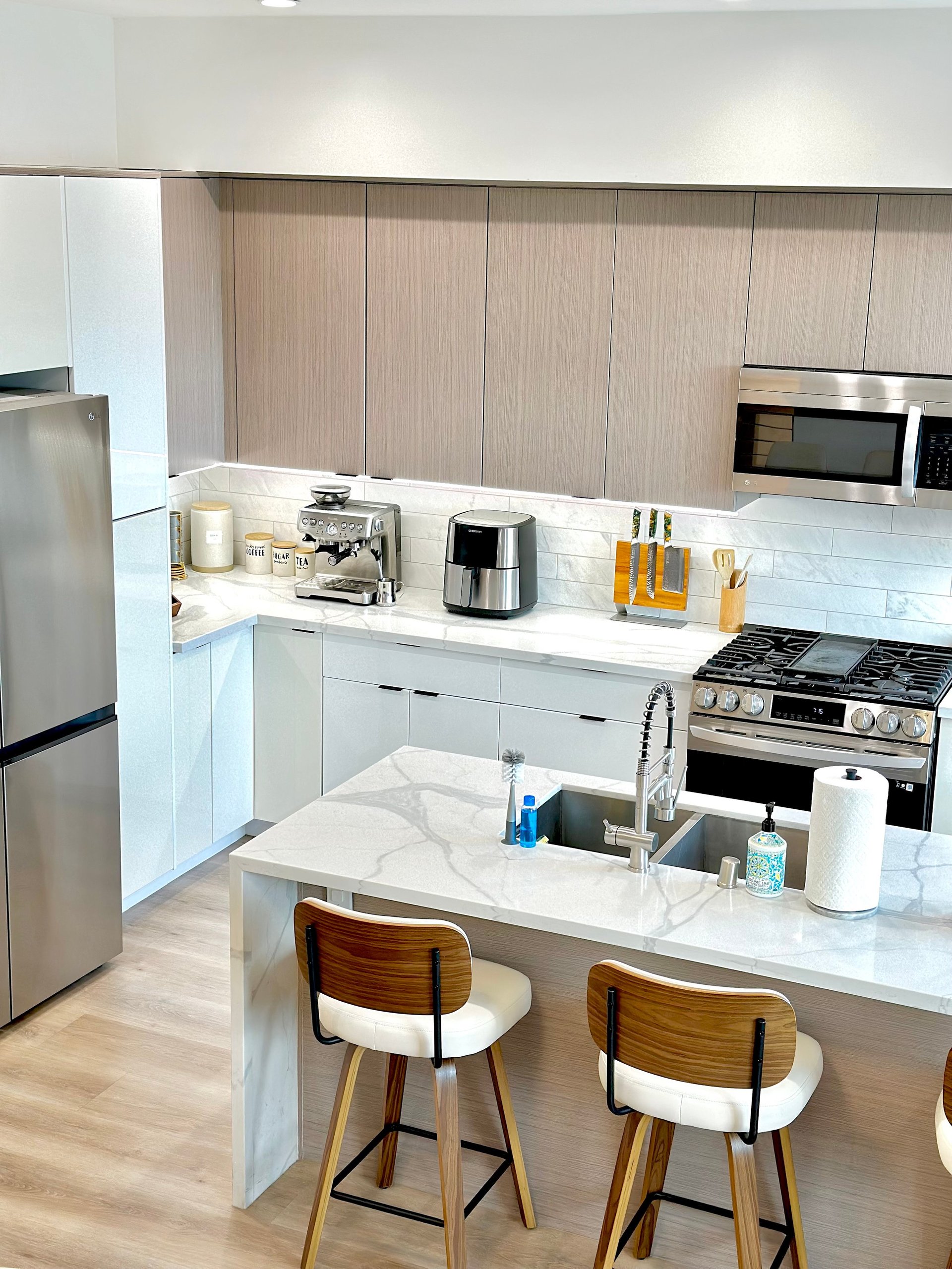Modern kitchen with white cabinetry, light wood backsplash, stainless steel appliances, white marble island with two wooden bar stools, and wood flooring.