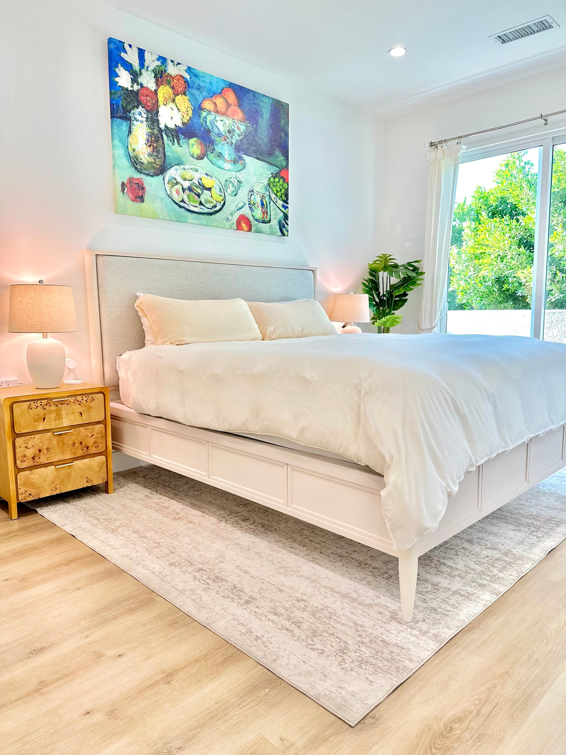 Bright modern bedroom with white bed, gray headboard, wooden nightstand, colorful fruit artwork, and large windows overlooking greenery