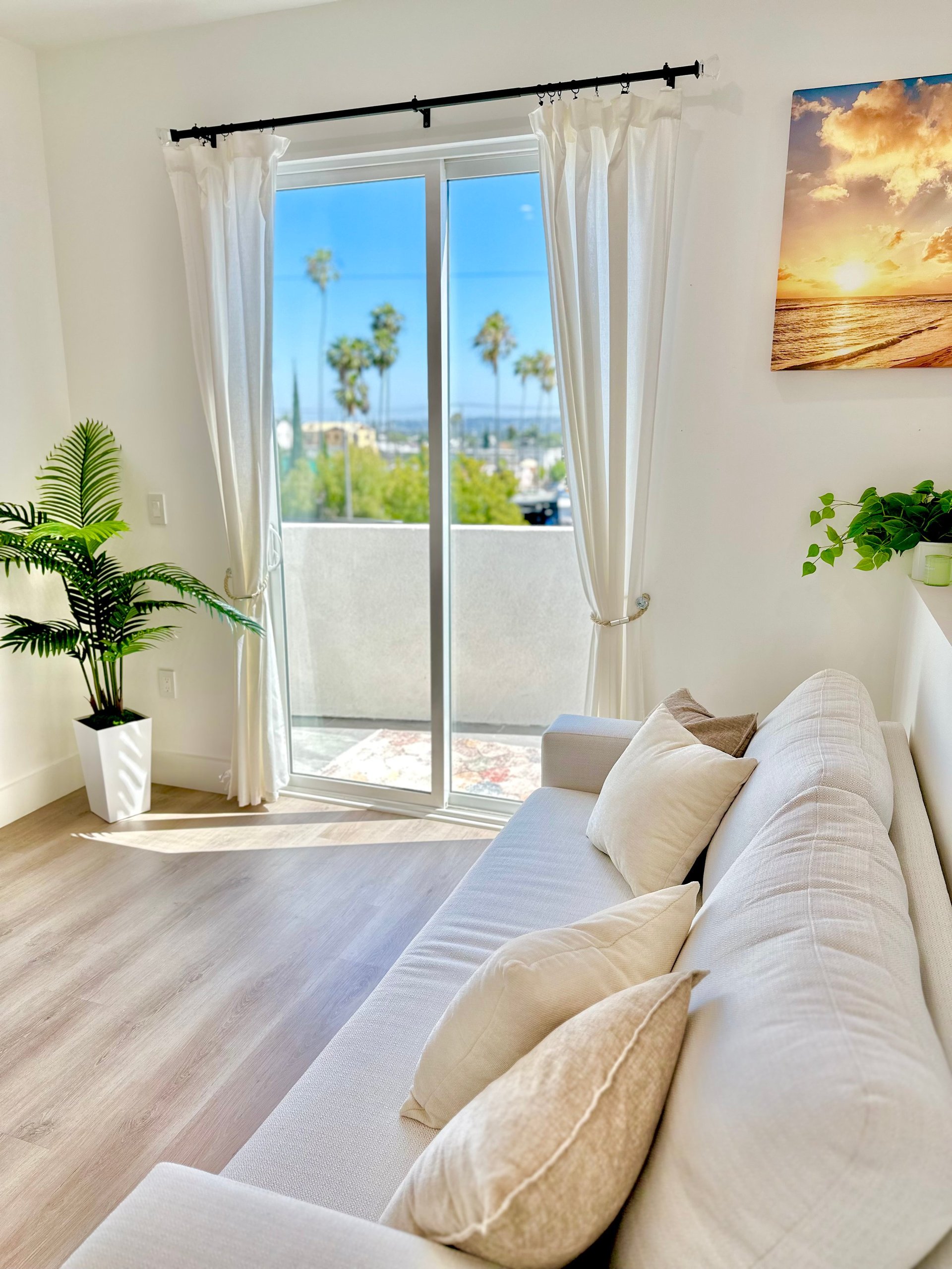 Bright bedroom with white bed, cream curtains framing glass doors opening to city view, plants on windowsill, and sunset artwork on wall