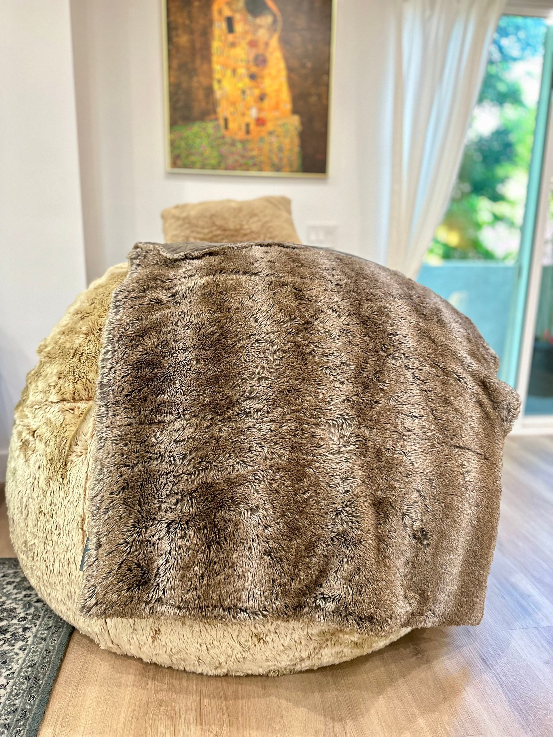 Round beige textured pouf in modern living room with artwork on wall and glass doors overlooking garden