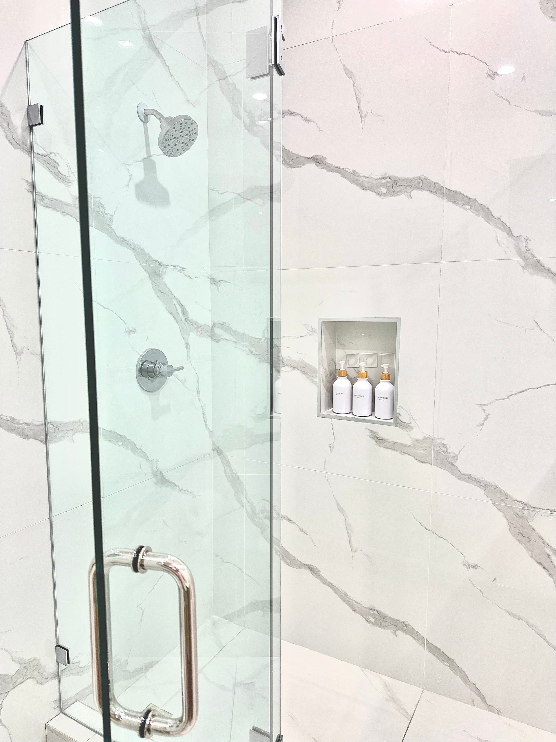 Modern glass shower enclosure with marble tile walls, chrome fixtures, and recessed niche holding white bottles