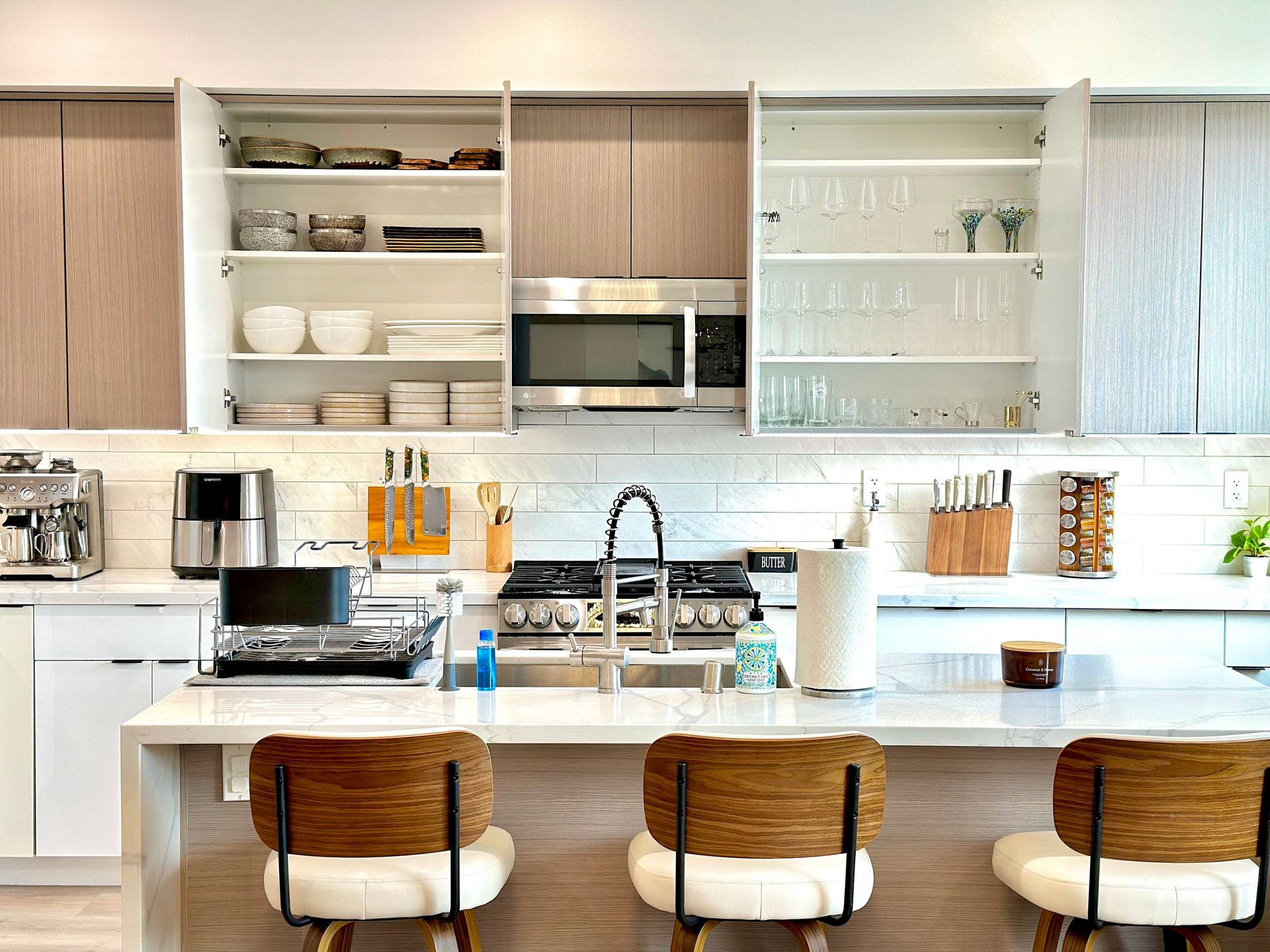 Modern kitchen with white counters, wooden bar seating, stainless steel appliances, and open shelving with dishware and glassware