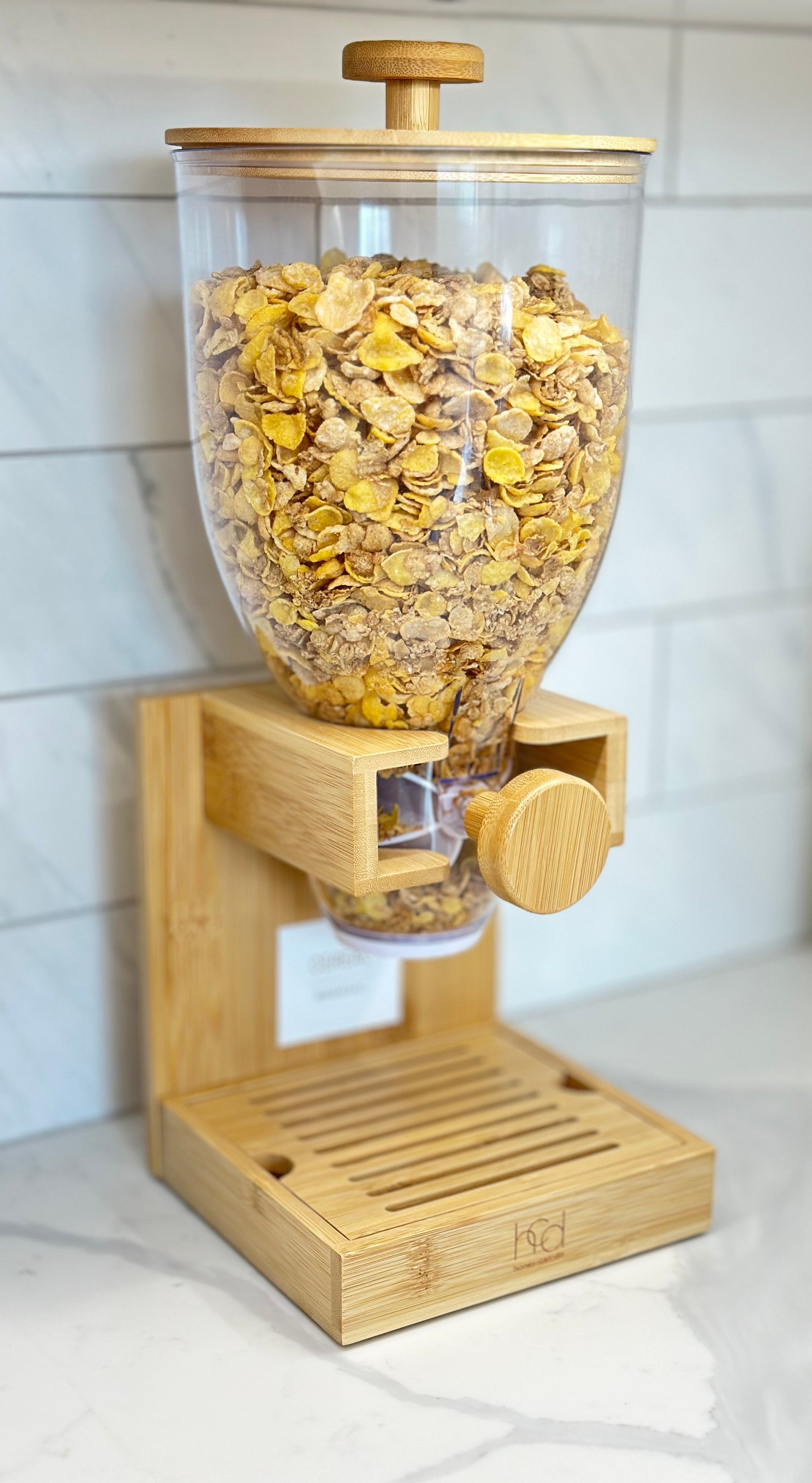 Wooden cereal dispenser with glass container filled with cornflakes and a control knob, mounted on a catchment tray