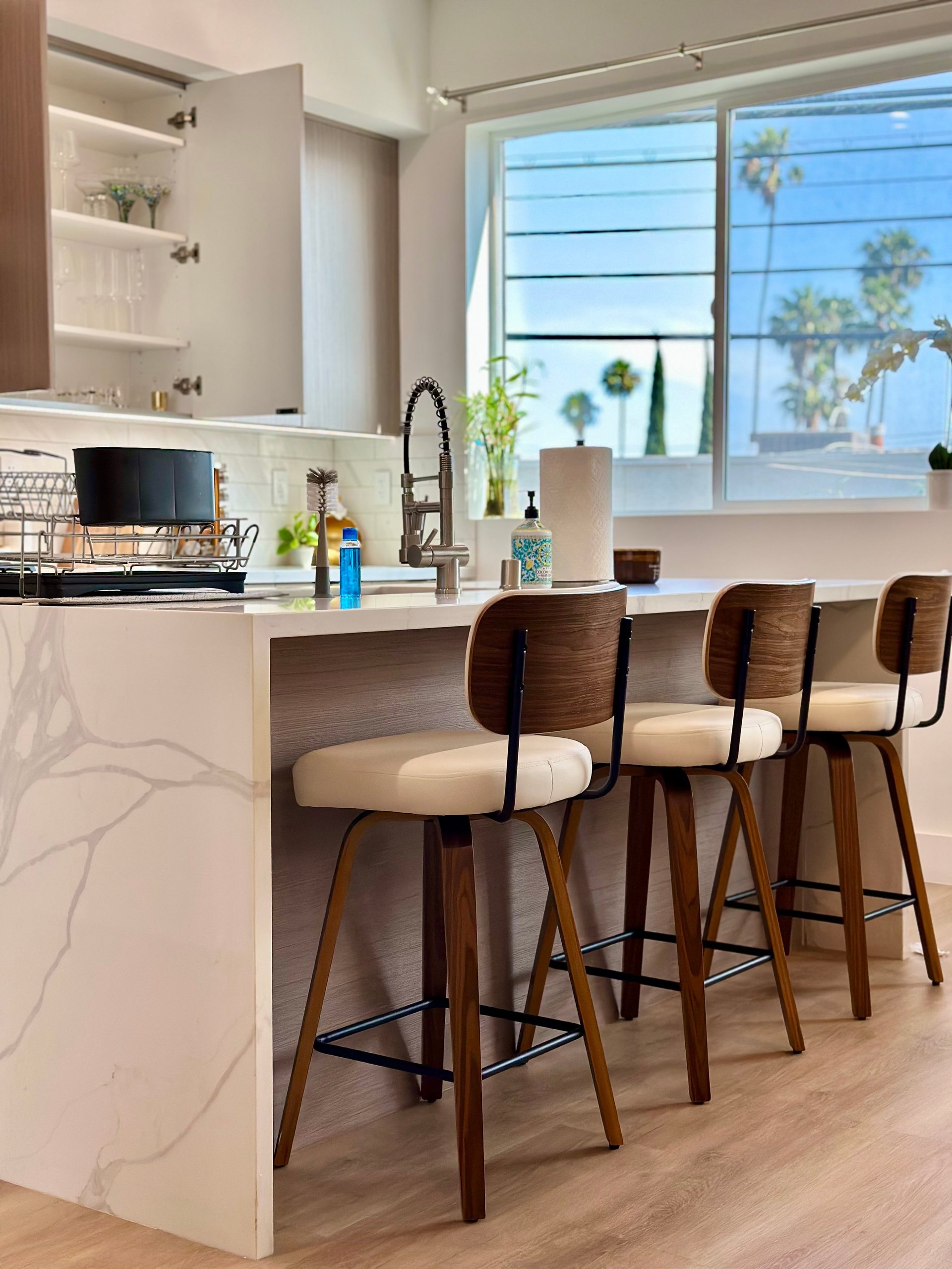 Modern kitchen with white island, three wooden bar stools, stainless steel appliances, and large windows overlooking palm trees