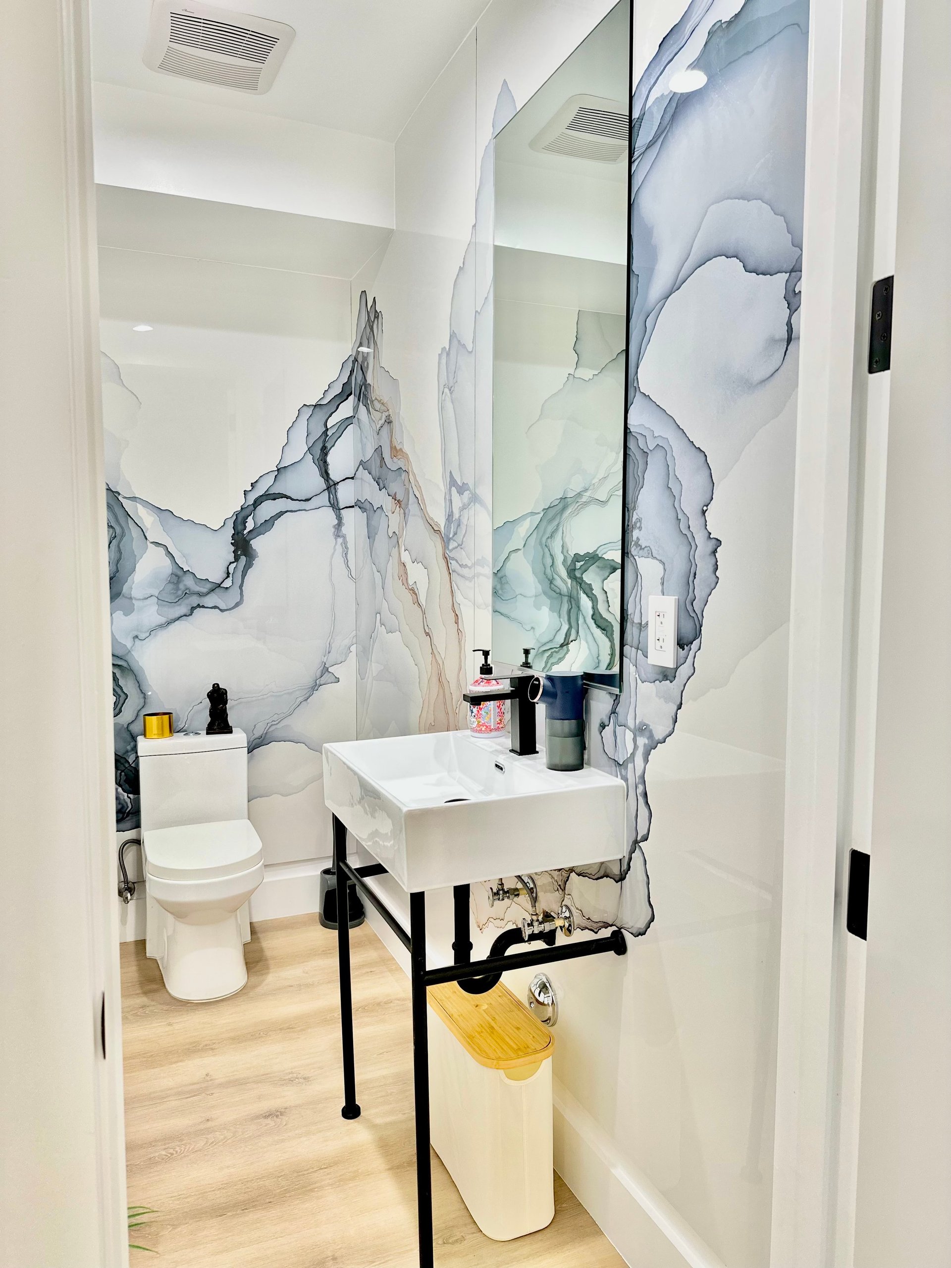 Modern bathroom with white sink on black stand, marble-patterned wall tiles, toilet, and decorative accessories on light wood flooring