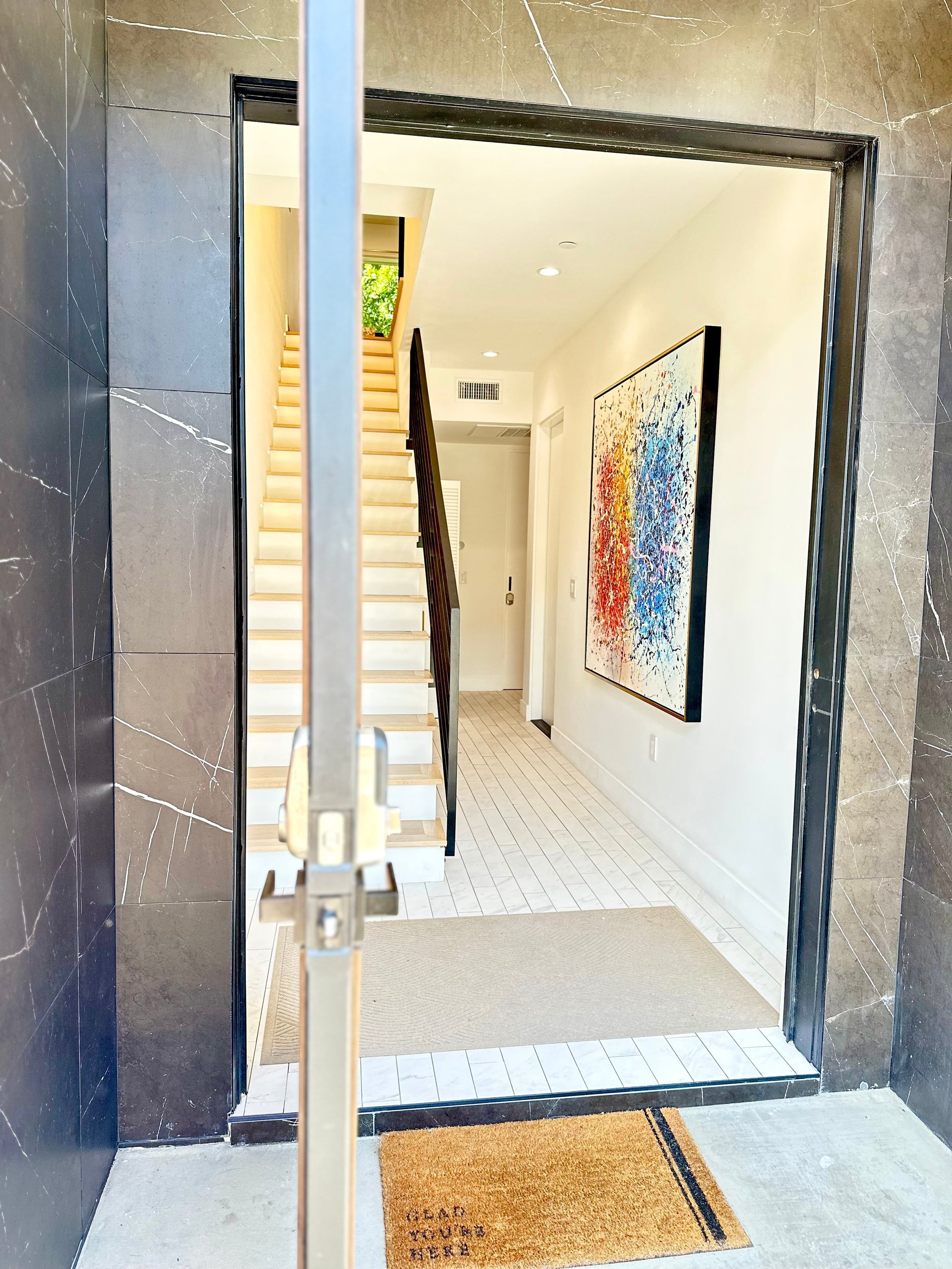 View through glass doors into a modern entryway with white flooring, stairs, and abstract artwork on the wall.