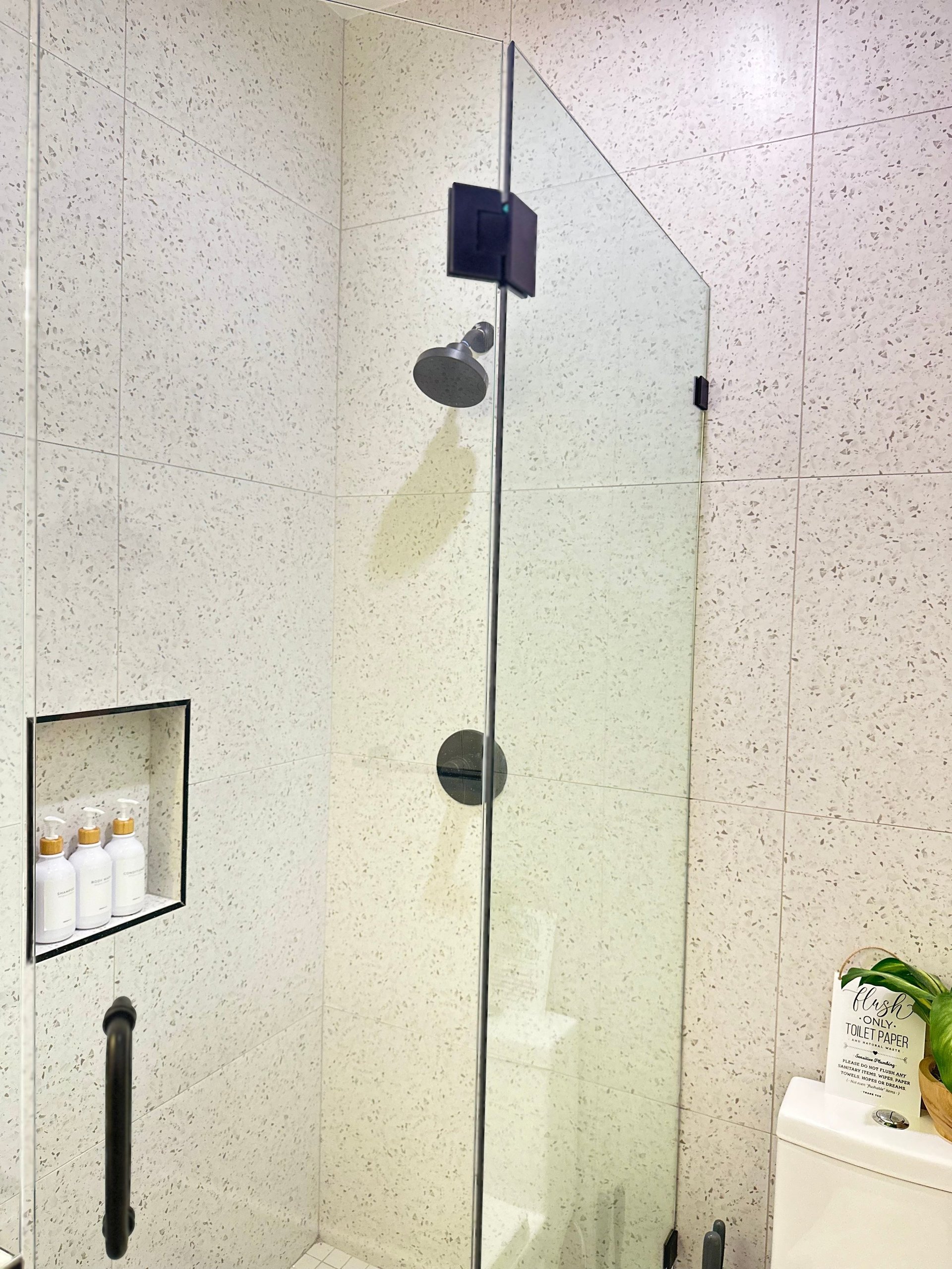Modern shower enclosure with frameless glass door, speckled beige tile walls, chrome fixtures, and a small recessed shelf with amenities