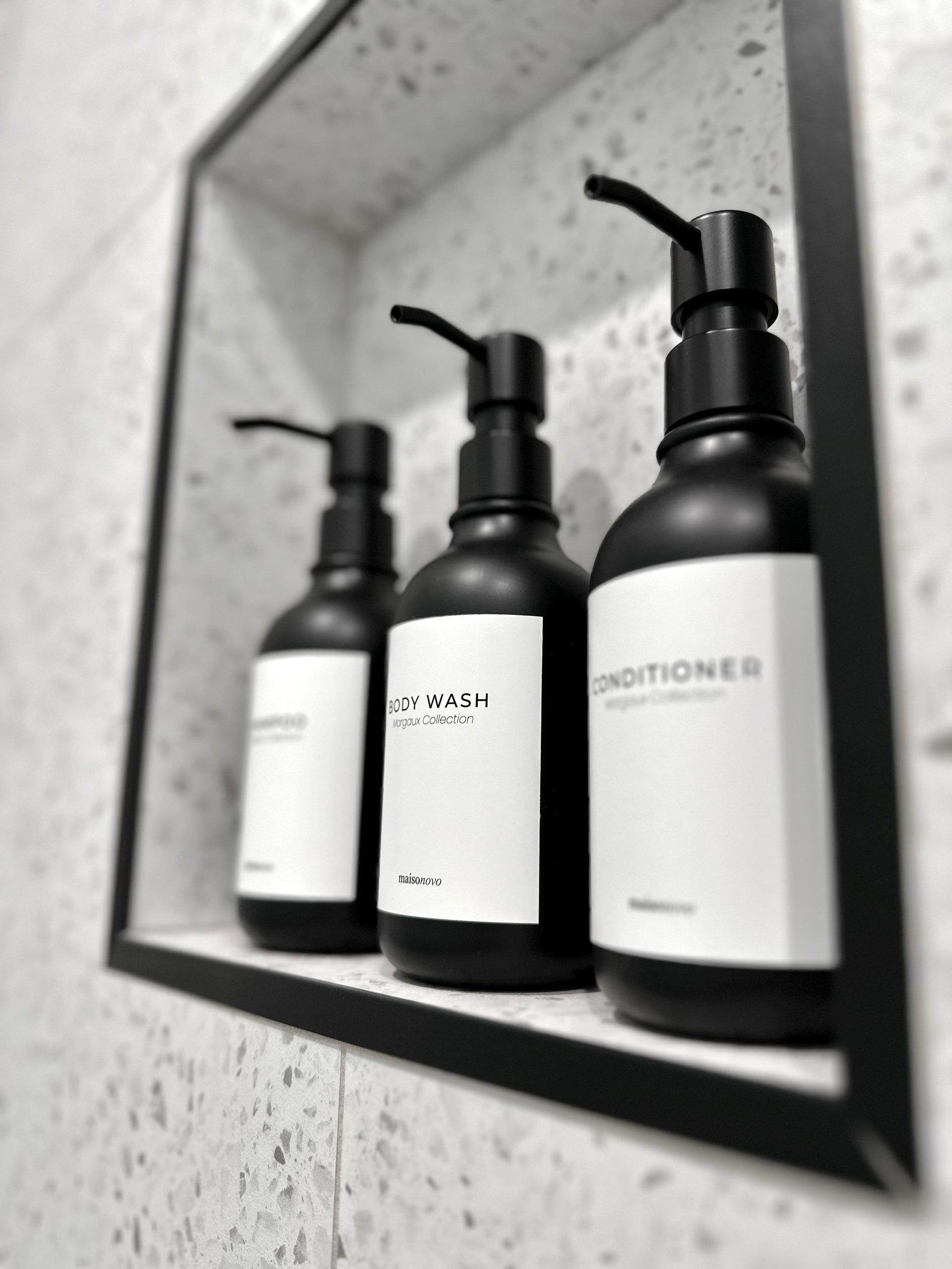 Three black and white pump bottles labeled Body Wash and Conditioner displayed in a recessed bathroom shelf with concrete walls.