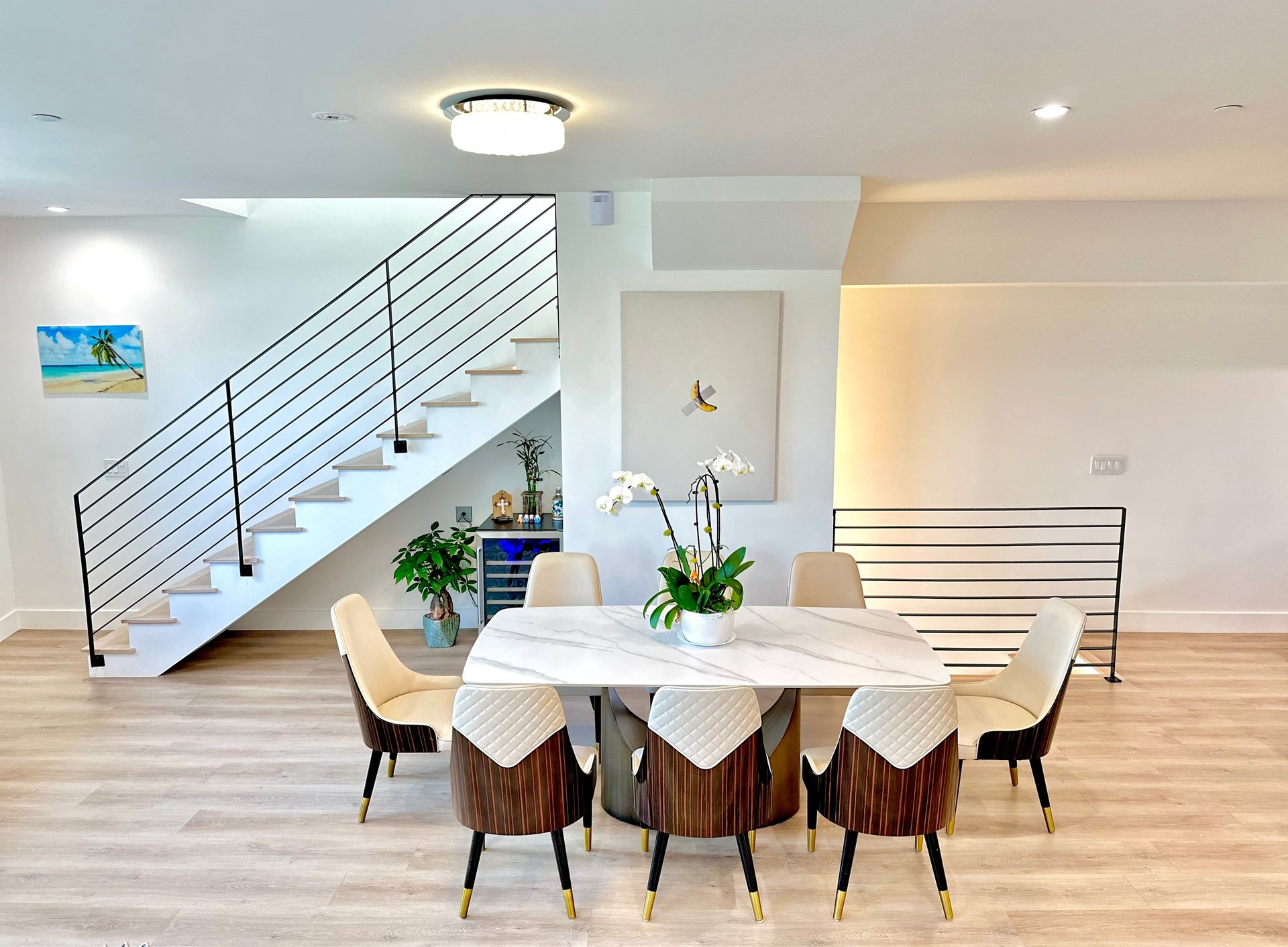 Modern dining room with round white table, cream chairs, wooden floor, and staircase with metal railings in bright minimalist interior